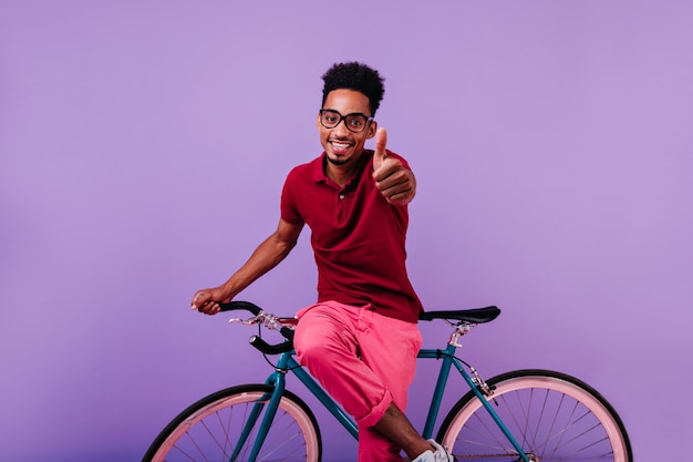 엄지 손가락으로 포즈를 취하는 안경에 영감을 된 아프리카 남자. 자전거에 앉아 웃 고 자신감 흑인 남자.