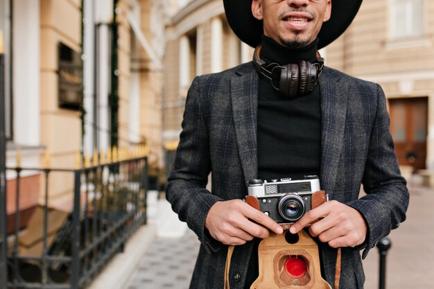 Inspired african man wears black shirt standing on street with camera in hands. Outdoor shot of well-dressed photographer.