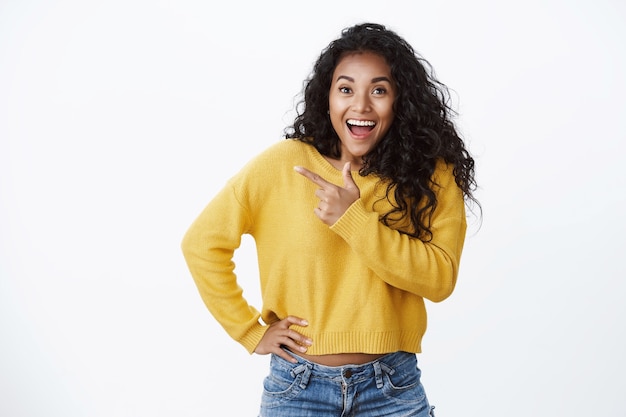 Inspired amused african-american woman yellow sweater gasping shocked and astonished, cheering emotionally telling great news, hold hand waist, pointing left blank space, standing white wall