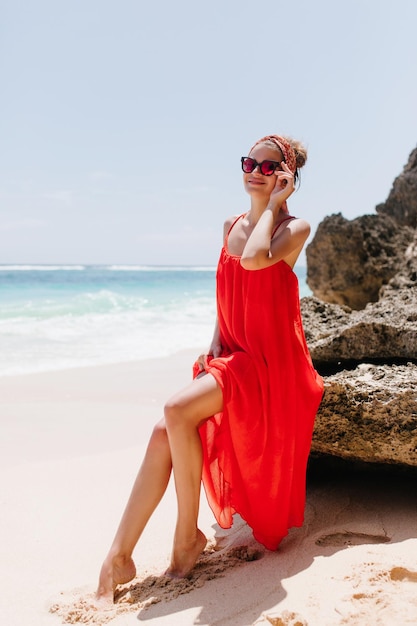 Inspired barefooted woman having fun at wild beach Outdoor photo of fascinating tanned female model in red dress expressing happiness on sea background