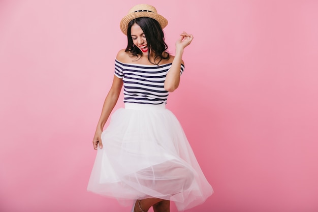 Inspired black-haired girl in straw hat dancing and singing. Indoor photo of magnificent latin woman in lush skirt.