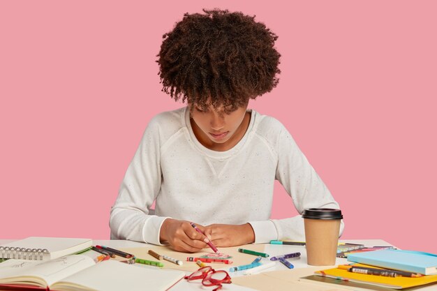 Inspired black young woman enjoys drawing with crayons on blank sheet of paper, focused down, has mood for creativity, creats something original, sits at workplace alone against pink wall