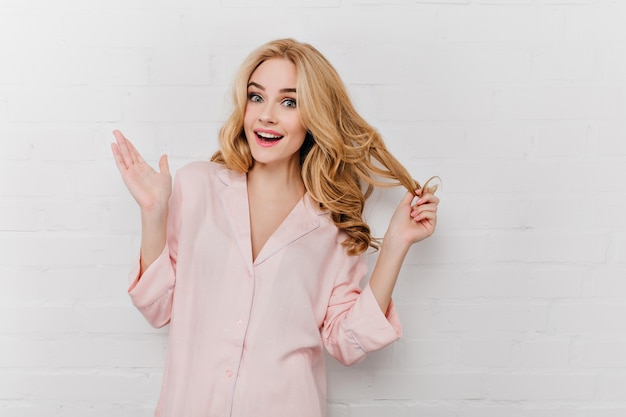 Inspired blue-eyed female model plays with her wavy hair. Portrait of good-looking girl in pink pajamas having fun in morning.