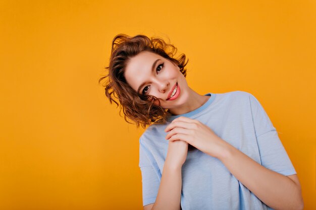 Inspired brunette girl with pink lips posing with pleasure on yellow wall. Studio shot of adorable caucasian woman in blue attire.