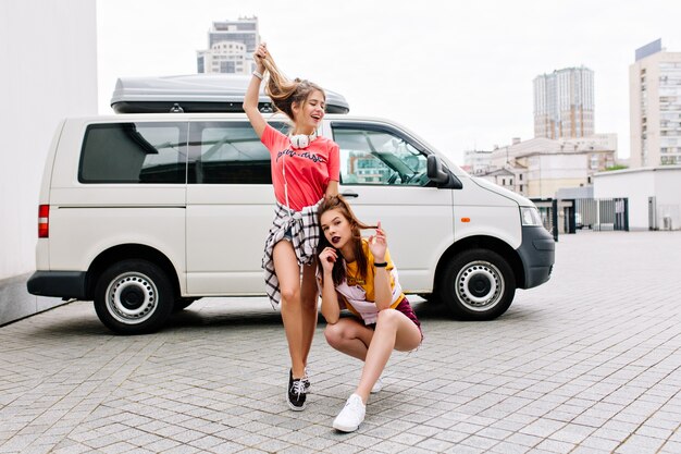 Inspired brunette girl in yellow shirt sits with legs crossed beside white car while her friend playing with long hair