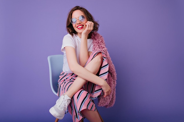 Inspired caucasian lady in white tshirt and shoes dreamy posing on chair Smiling charming girl with wavy hair sitting on purple background