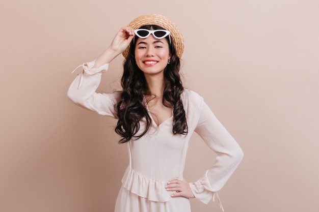 Inspired curly korean woman smiling at camera. Front view of romantic asian young woman in sunglasses isolated on beige background.