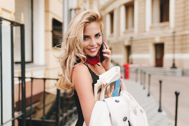 Inspired curly woman holding jacket laughing while speaking with someone on phone