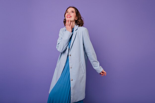 Inspired female model in long blue coat laughing  Portrait of smiling gorgeous girl with curly hairstyle wears autumn outfit.