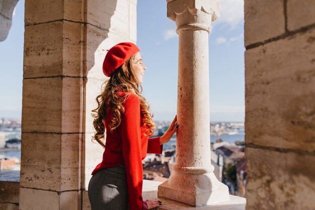 Inspired french girl with curly hairstyle standing near stone columns and enjoying city views