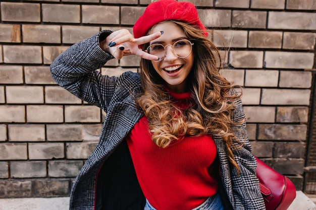 Free photo inspired french lady with elegant hairstyle posing with peace sign on brick wall background