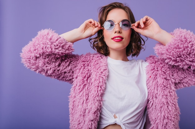 Foto gratuita ragazza ispirata in abito luminoso in posa sulla parete viola. carina giovane donna con i capelli ricci indossa soffice giacca di pelliccia.