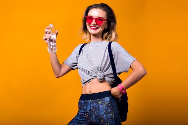 Inspired girl in red sunglasses with short hairstyle holding bottle of water and smiling. Indoor portrait of laughing european female model isolated on yellow background.