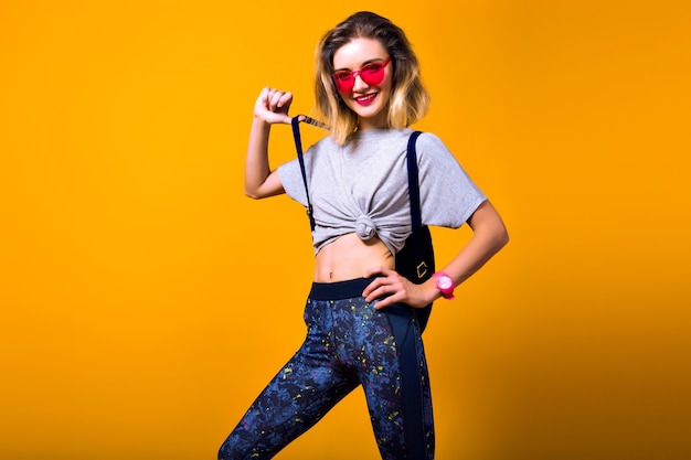 Inspired girl in red sunglasses with short hairstyle holding bottle of water and smiling. indoor portrait of laughing european female model isolated on yellow background.