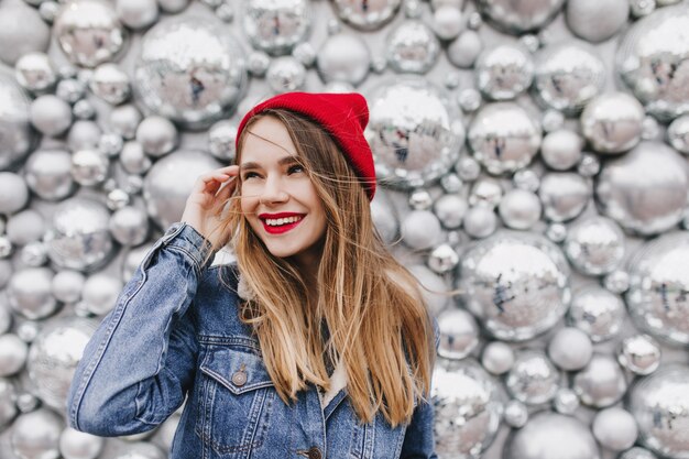Вдохновленная девушка с прямыми каштановыми волосами смотрит в сторону с улыбкой во время фотосессии с праздничными аксессуарами. Фото симпатичной европейской женщины в красной шляпе, стоящей возле дискотечных шаров.