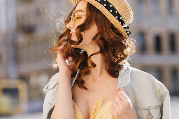 Inspired girl with shiny red hair posing on blur street background Outdoor photo of magnificent ginger woman in stylish hat