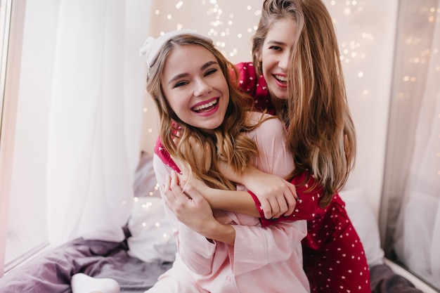 Inspired girl with sincere smile embracing friend in morning. Positive sisters with long hair hugs in bedroom.