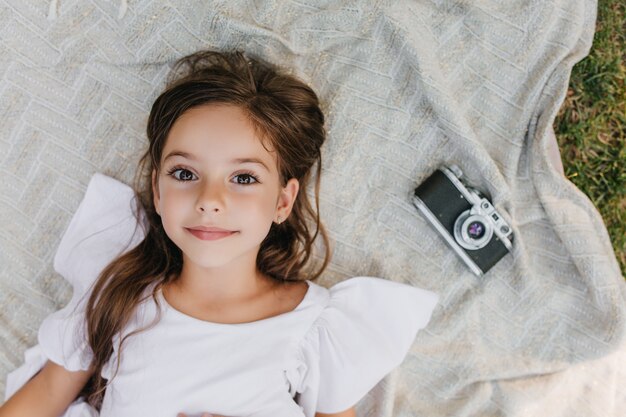 Inspired little lady with big brown eyes lying on blanket in garden and looking up with gently smile. Overhead portrait of dark-haired girl in white dress relaxing on the ground near camera.