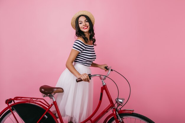 Inspired shapely woman standing with bicycle and looking away. Blissful brown-haired girl in hat enjoying indoor photoshoot.