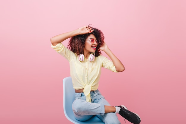 Inspired slim girl in vintage yellow shirt stretching on chair. Indoor portrait of lovely curly african lady in black sneakers looking away with smile.