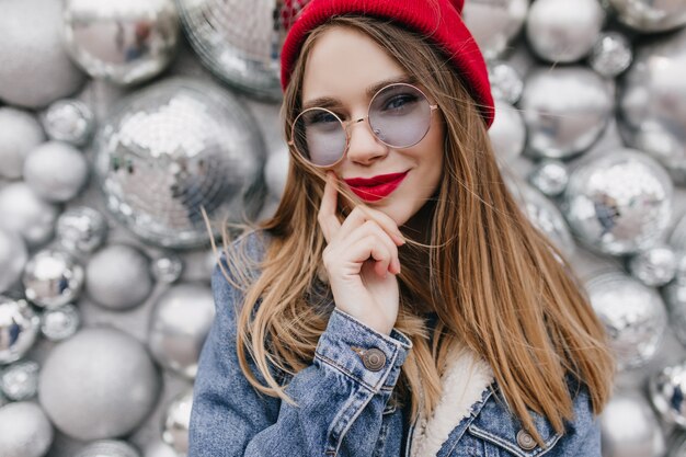 Inspired white female model touching her face while posing on sparkle wall. Smiling fascinating woman in round glasses and denim jacket standing in front of disco balls.