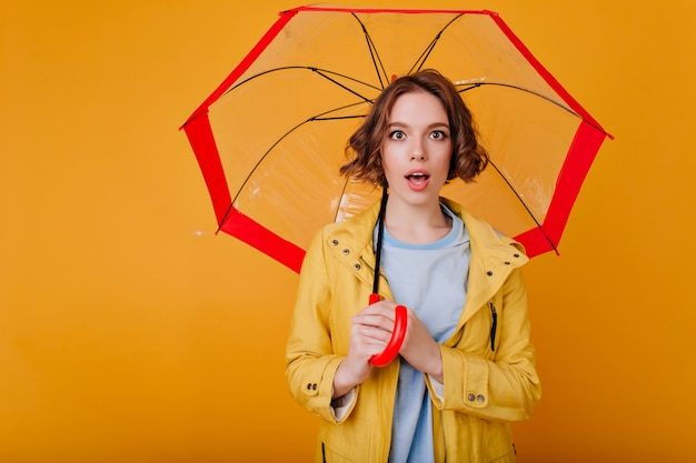 赤い日傘を手に黄色い壁に立っている驚きの表情を持つインスピレーションを得た白人の女の子。スタイリッシュな傘でポーズをとる秋の服装で夢のようなブルネットの女性の写真。