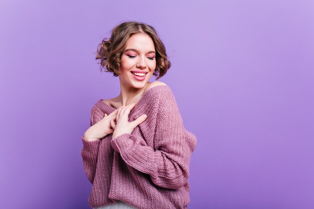 영감을 얻은 백인 여성은 수줍은 미소로 포즈를 취하는 부드러운 니트 스웨터를 입습니다. 유행 헤어 스타일 보라색 벽에 웃 고 열정적 인 젊은 아가씨의 실내 초상화.