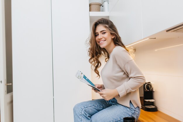 Inspired white young woman in beige shirt sitting on table in kitchen with newspaper