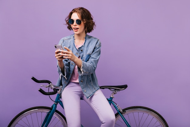 Free photo inspired woman in casual attire looking at phone screen. joyful white girl in dark sunglasses using smartphone with bicycle.