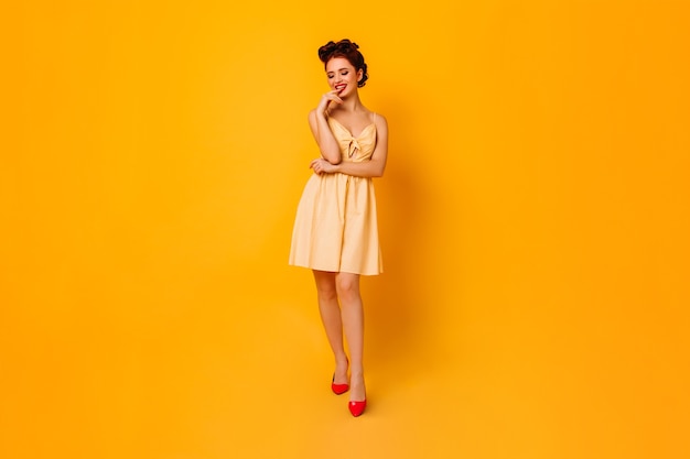 Inspired woman playfully posing on yellow space. Amazing pinup lady in short dress enjoying photoshoot.