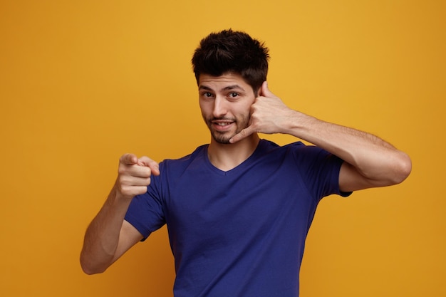 Inspired young handsome man looking at camera doing call me gesture pointing to camera on yellow background