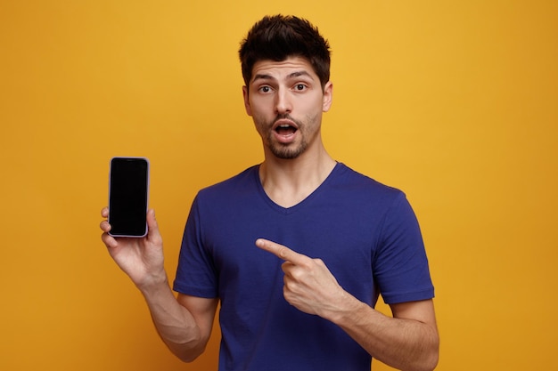 Inspired young handsome man looking at camera showing mobile phone pointing to it on yellow background