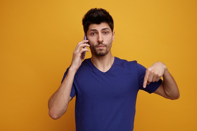 Inspired young handsome man talking on phone looking at camera pointing down on yellow background