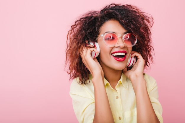 Inspired young lady with light-brown skin happy laughing while listening favorite song. Close-up catching mulatto woman in casual comfortable attire holding headphones.