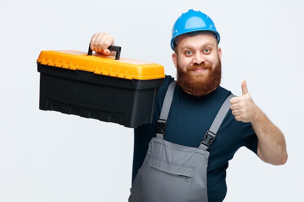 Free photo inspired young male construction worker wearing safety helmet and uniform raising tool box looking at camera showing thumb up isolated on white background