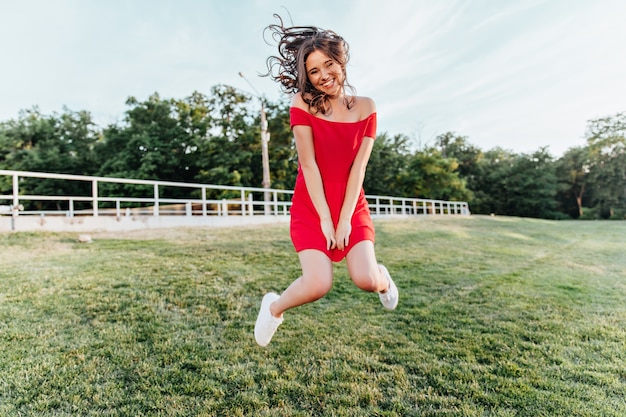 公園に飛び込んで笑っているインスピレーションを得た若い女性。夏の週末に楽しんでいる赤いドレスの美しいブルネットの少女。