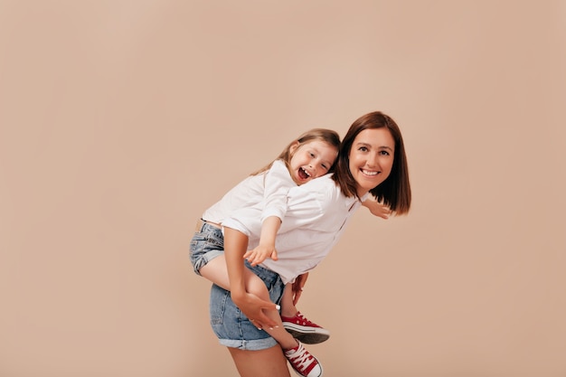 Inspired young woman without make-up spending time with daughter, carrying her piggyback over isolated background.