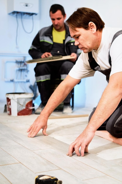 建設現場の建設作業員へのセラミック床タイルの設置セメント接着剤で床にタイルを敷設する作業員床の改修垂直フレーム