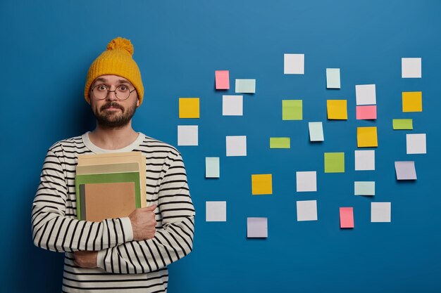 intelligent bearded student in yellow hat, striped jumper prepares for workshop, stands with papers and notepad