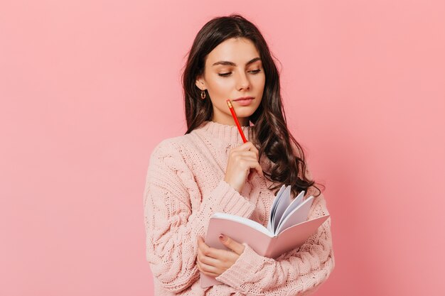 Умная женщина в розовом свитере думает о продолжении своей книги. Брюнетка смотрит в дневник на розовом фоне.