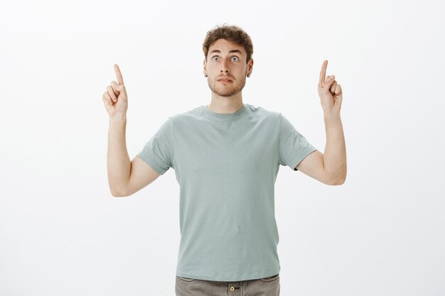 Intense afraid european guy with fair hair, pointing upwards with raised index fingers and staring aside on something scary and shocking