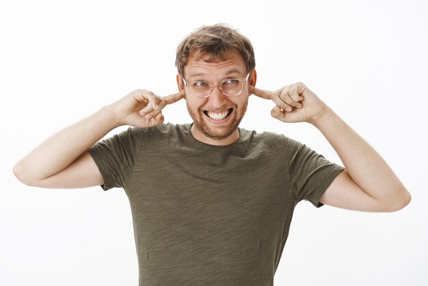 Intense male scinentist preparing for loud bang clenching teeth covering ears with index fingers and gazing with await at upper right corner in glasses and green t-shirt hearing noisy sound