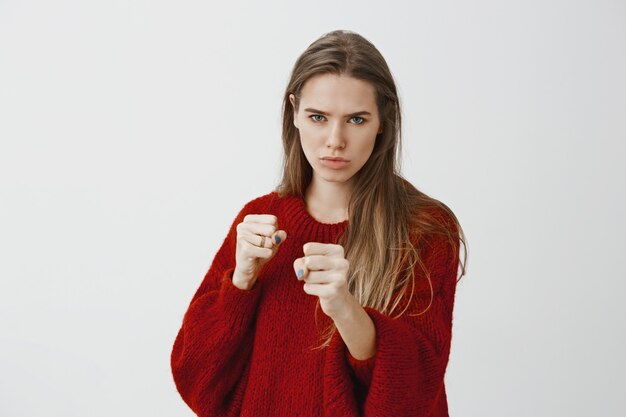 Intense serious woman ready to fight for love. Focused good-looking european female model in stylish red loose sweater, standing in boxing pose with raised clenched fists, frowning, ready to defense