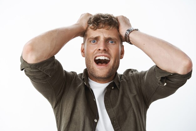 Intense shocked guy in stupor with blue eyes and fair hair holding hands on head yelling, screaming in panic, freaking out, standing stunned and insecure over gray wall scared and frightened