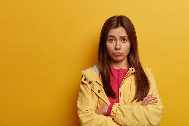 Intense upset woman keeps hands crossed, regrets missing interesting chance, frowns face, looks in dissatisfaction, wears pink jumper and yellow anorak