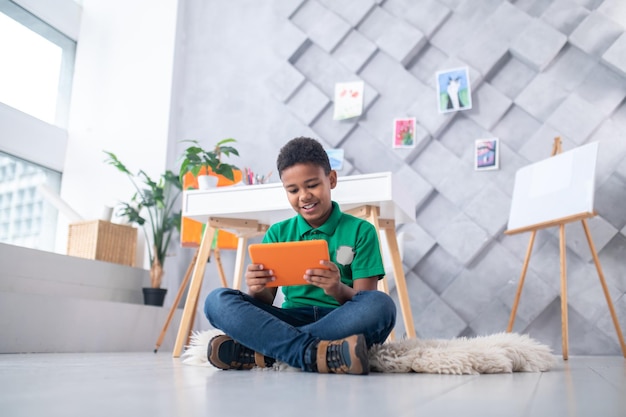 Interest. Smiling dark-skinned boy of school age in green t-shirt and jeans sitting on floor looking with interest at tablet in bright room with easel and paintings