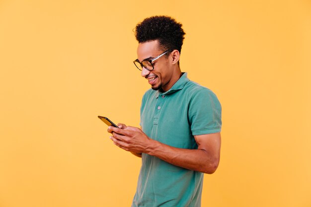 陽気な笑顔で電話の画面を見ている興味のある黒人男性。メッセージを読んでメガネでハンサムなアフリカの少年の屋内ショット。