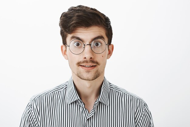 Interested bookworm wants buy new book in store. Portrait of excited timid european male model with moustache and beard in glasses, lifting eyebrows from surprise, listening carefully over gray wall