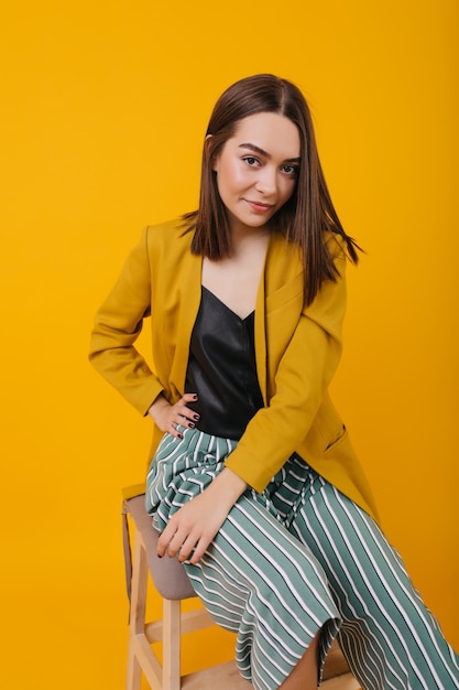 Free photo interested brunette girl in bright jacket sitting on chair. lovely brown-haired woman in striped pants posing with pleasure.