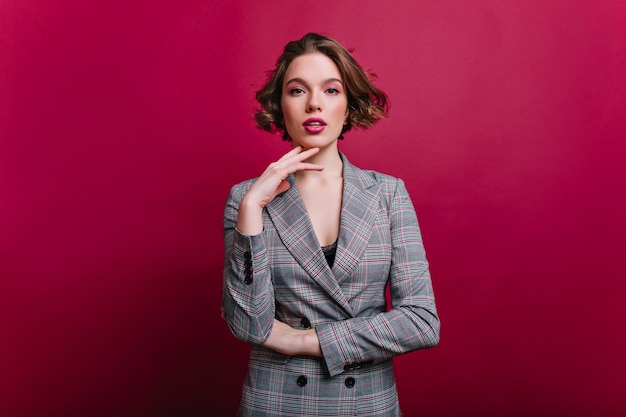 Interested businesswoman with trendy makeup posing on claret wall. Indoor photo of serious young lady in tweed jacket standing in confident pose.